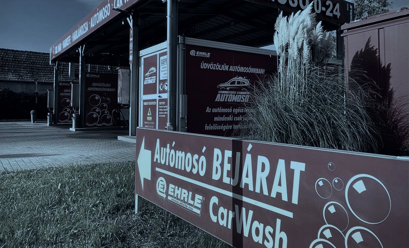 A telephely bejáratát jelző tábla a fűben, mögötte a fedett mosóállások sora.