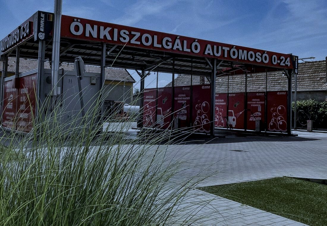 A telephely kapuja délután, az ÖNKISZOLGÁLÓ AUTÓMOSÓ 0–24 felirattal, mögötte a fedett mosóállások.