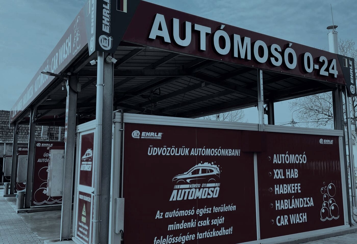 A telephely fedett mosóállása az AUTÓMOSÓ 0–24 felirattal, mellette a szolgáltatásokat felsoroló piros tábla.